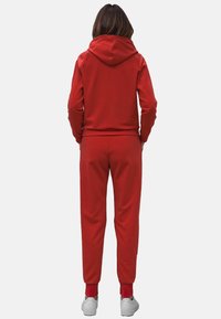 Felpa rossa con cappuccio e pantaloni coordinati, realizzati in tessuto morbido. Caratteristiche includono un cappuccio, polsini e caviglie a costine, abbinati a sneaker bianche. Design semplice.