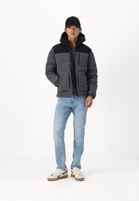 Grå pufferjakke med sorte detaljer og hætte, med lynlåslomme foran, båret over en sort skjorte og lyseblå jeans, parret med sneakers.