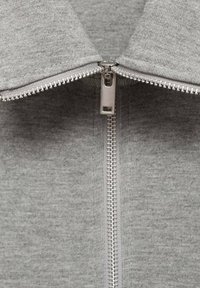 Grijze zip-up sweatshirt van een textuurstof, met een zilveren rits en een hoge kraag. De rits loopt verticaal over het midden.