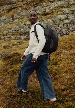 Mann iført hvit genser, blå jeans og svart ryggsekk som går tur over steinete, gresskledt terreng og ser over skulderen.