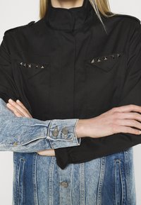 Černá bunda s ramenními epauletami a stříbrnými hřebíčky na hrudních kapsách, kombinovaná s rukávy z světle modrého denimu a knoflíkovými manžetami.