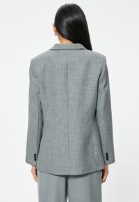 Blazer gris texturizado con un corte entallado, cuello con muescas y puños abotonados. La tela presenta un sutil patrón de espiga.