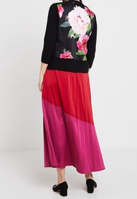 Kobieta w czarnej bluzce z dużym różowym i czerwonym wzorem kwiatowym na plecach oraz długiej plisowanej spódnicy w kolorze czerwonym i różowym, w czarnych szpilkach.