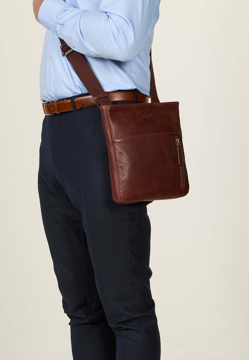Borsa a tracolla in pelle marrone con tasca frontale con zip, tracolla regolabile e texture liscia, indossata di lato.