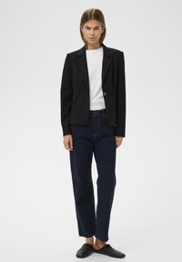 Fekete, szabott blézer egy gombbal, fehér pólóra véve, sötétkék, bő szárú farmernadrággal és fekete slip-on cipővel párosítva.