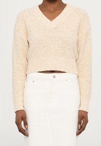 Personne portant un pull court texturé en maille crème à col en V et un pantalon blanc taille haute, les mains détendues le long du corps.