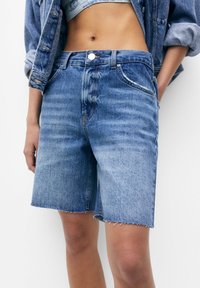 Pantalones cortos de mezclilla en azul claro con dobladillo deshilachado, diseño de cinco bolsillos y cierre de botón. Acompañados de una chaqueta de mezclilla a juego.