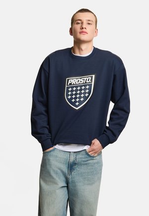 Młody mężczyzna w granatowej bluzie z logo "PROSTO." i jasnoniebieskich jeansach, stojący z rękami w kieszeniach na białym tle.