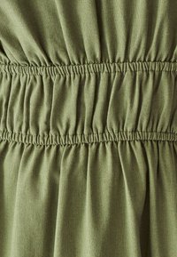 Tessuto verde oliva con un aspetto texture, caratterizzato da gather elastici in vita. Materiale morbido e drappeggiato con una leggera lucentezza.