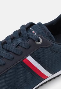 Marinblå textilsko med en texturerad yta, platta snören, ett vitt och rött randigt detalj och en silverfärgad ögla.