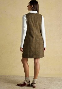 Braun karierter Dress mit taillierter Passform, getragen über einem weißen Langarmshirt, kombiniert mit bordeauxroten Schuhen und beigen Socken.