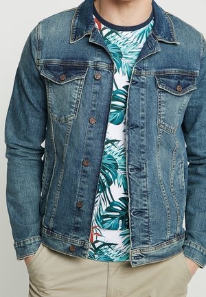 Persona che indossa una giacca di jeans blu sopra una t-shirt bianca con motivi di foglie tropicali verdi e pantaloni beige, con le mani nelle tasche.