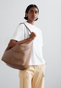 Sac hobo en cuir tan avec une finition lisse, une ligne supérieure courbée et un design minimaliste. Marquage doré visible à l'avant.