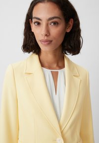 Yellow blazer with notched lapels, soft fabric, single button closure, paired with a white top featuring a keyhole design.