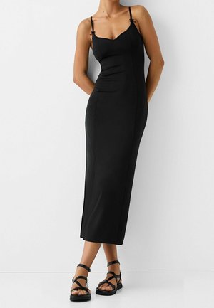 Kobieta w dopasowanej czarnej midi sukience na ramiączkach typu spaghetti bez rękawów, w czarnych płaskich sandałach na paski, stojąca przy białej ścianie.