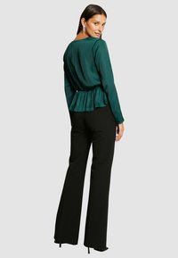 Haut en satin vert à manches longues et ourlet en peplum, associé à un pantalon noir à jambes larges. Modèle de dos, portant des boucles d'oreilles accrocheuses.