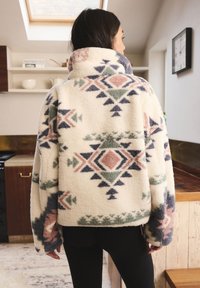 Giacca in pile con collo alto, caratterizzata da una base color crema e motivi geometrici in tonalità di blu, verde e rosa. Silhouette oversize.