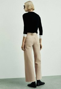 Top nero a maniche lunghe, pantaloni beige a gamba larga, tessuto testurizzato, due tasche posteriori, indossato con stivaletti neri, su uno sfondo chiaro.