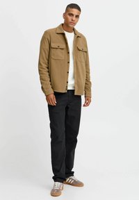 Bruine fleece jas met twee borstzakken, knoopsluiting, gedragen over een witte trui, gecombineerd met zwarte broek en grijze sneakers.