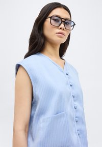 Νέα γυναίκα που φορά μεγάλα μαύρα γυαλιά και αμάνικο γαλάζιο μπλουζάκι με ρίγες και κουμπιά καλυμμένα με ύφασμα.