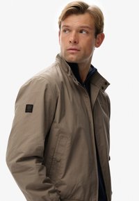 Beige lichtgewicht jas met een staande kraag, ritssluiting en zijzakken. Voorzien van een logo-opdruk op de linker mouw en een gladde textuur.