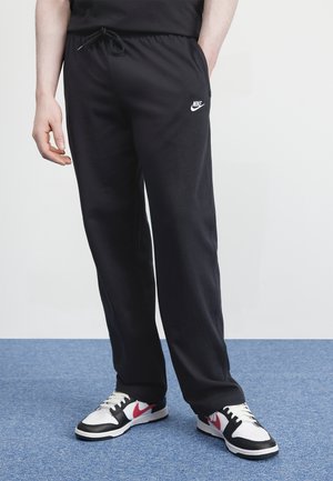 Man draagt zwarte Nike joggingbroek met witte en rode Nike sneakers, staand op een blauw tapijt tegen een witte muur.