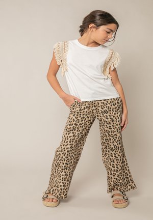 Jeune fille posant en haut blanc à franges sans manches, pantalon imprimé léopard, et sandales beige sur fond beige.