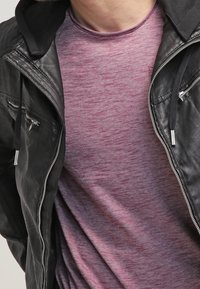 Musta nahkatakki, jossa on hopeiset vetoketjut, marjapunaiseen, teksturoituun T-paitaan päällä. Takissa on huppu, mikä tuo lisäkerroksia ja yksityiskohtia.