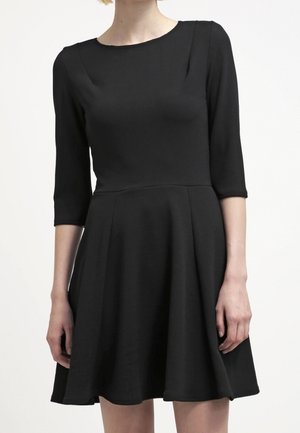 Kvinde iført en sort knælængde kjole med trekvart ærmer og en figursyet overdel, stående foran en ensartet hvid baggrund.