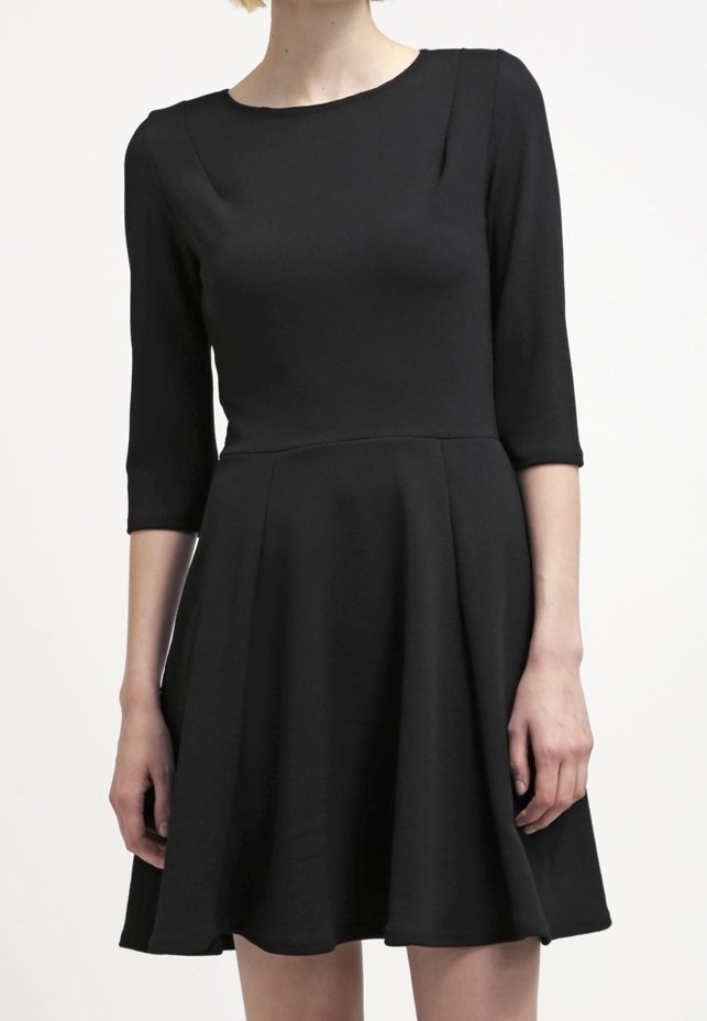 Femme portant une robe noire longueur genou avec des manches trois-quarts et un corsage ajusté, debout devant un fond blanc uni.
