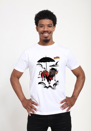 Man smiling with hands on hips wearing a white t-shirt featuring a cartoon dog in red suit holding a black umbrella and flying among clouds.