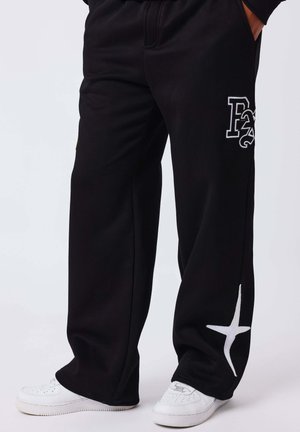 Personne portant un pantalon de survêtement noir avec des motifs graphiques blancs près de la cuisse et du bas de la jambe, associé à des baskets blanches.