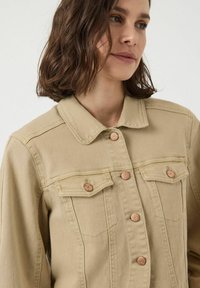 Een vrouw met bruin haar draagt een beige denimjack met koperen knopen, terwijl ze iets naar links kijkt tegen een effen lichte achtergrond.