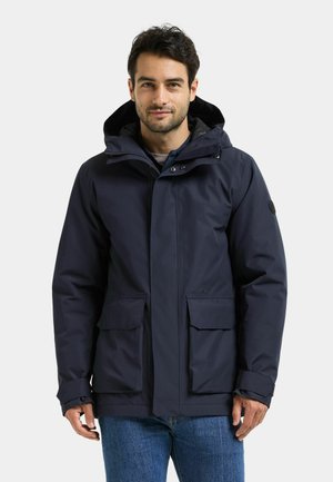 Mand iført en mørk navy hættetrøje med forlommer og blå jeans, stående mod en ensfarvet lys baggrund.
