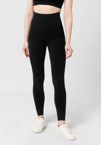 Zwarte, high-waisted legging gemaakt van een rekbaar materiaal, met een gladde textuur en een strakke pasvorm, gecombineerd met witte sneakers.