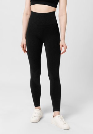 Leggings de tiro alto negros fabricados con un material elástico, que presentan una textura suave y una silueta ajustada, combinados con zapatillas blancas.