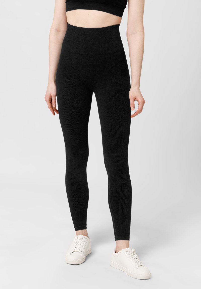 Zwarte, high-waisted legging gemaakt van een rekbaar materiaal, met een gladde textuur en een strakke pasvorm, gecombineerd met witte sneakers.