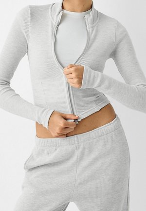 Veste zippée gris côtelé avec un col haut, associée à un pantalon de sport assorti. Le tissu semble doux et texturé. Détail de fermeture éclair visible.