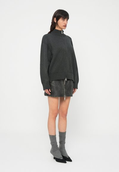 Camisola cinzenta de malha com fecho éclair e gola alta, combinada com uma mini saia de couro preta e meias cinzentas caneladas. Saltos pretos com bico afilado completam o conjunto.