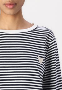 Maglietta a righe blu navy e bianca con un colletto a coste. Presenta un piccolo cuore beige sulla parte sinistra del petto.