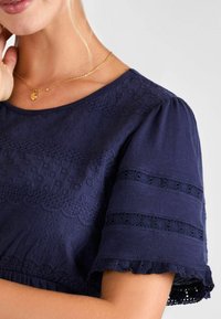 Tengerés kék, rövid ujjú felső, textúrált hímzéssel, hullámos szegéllyel és csipke részletekkel a szabott ujjain, kerek nyakkal.