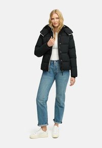 Chaqueta de plumas negra con capucha, que cuenta con un cierre de cremallera y botones a presión. Se lleva con jeans de lavado claro, ligeramente remangados, y zapatillas blancas.