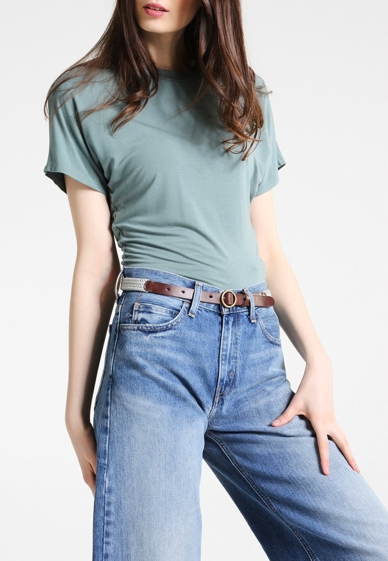 Femme portant une chemise vert clair à manches courtes rentrée dans un jean bleu taille haute avec une ceinture marron, debout devant un fond uni.
