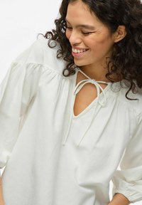 Hvit skjorte med lange ermer, V-hals og knyting ved halsen. Har innsamlede ermer og en avslappet passform. Myk tekstur.
