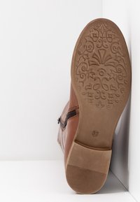 Semelle d'une botte en cuir marron avec un motif floral complexe sur la bande de roulement, taille 37 indiquée, reposant contre un coin de mur et de sol blanc.