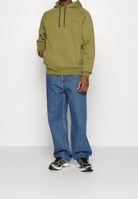 Persona con sudadera con capucha de color verde oliva con cordones negros, vaqueros azules holgados y zapatillas de running negras, blancas y plateadas, de pie frente a un fondo liso.