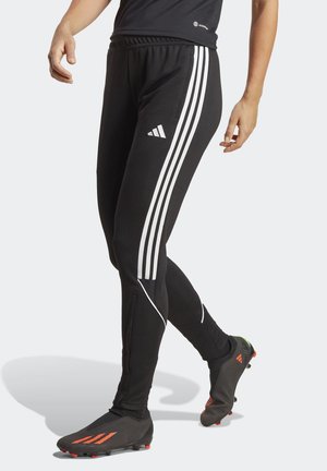 Jambes portant un pantalon de sport Adidas noir avec des bandes blanches et des chaussures de football noires avec des accents orange sur un fond gris clair.
