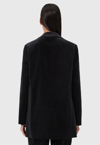 Donna con lunghi capelli scuri che indossa un blazer nero aderente, vista di spalle su uno sfondo grigio chiaro uniforme.