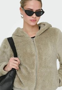 Flauschige beige Zip-Hoodie mit Kapuze, mit einer strukturierten Oberfläche und Fronttaschen. Modell trägt katzenaugenförmige schwarze Sonnenbrille und hält eine schwarze Tasche.