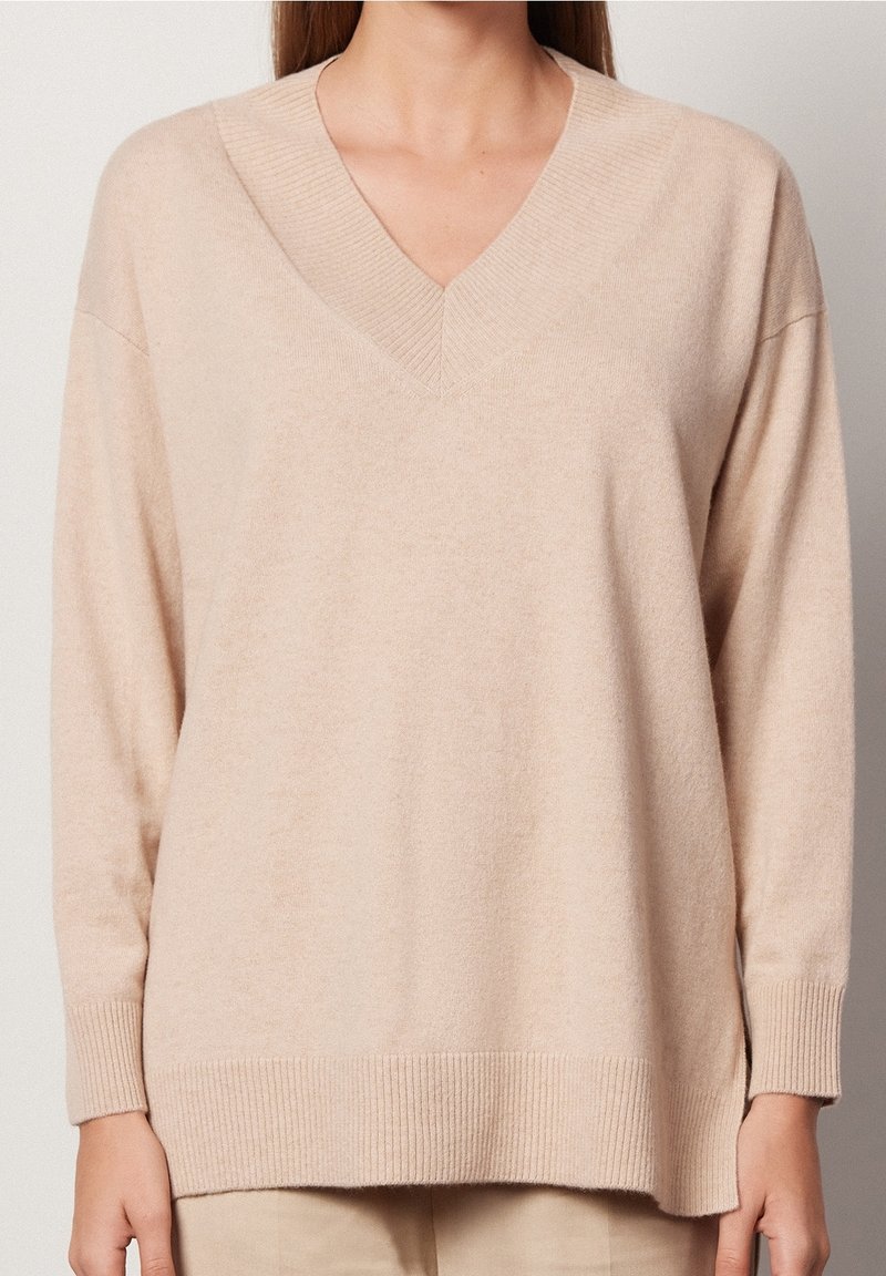 Pull beige à col en V avec manches longues, poignets et ourlet côtelés. Tissu doux et léger avec une coupe décontractée et des fentes latérales.